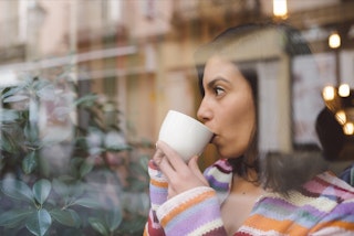 A woman looks out of a window while sipping from a white coffee mug. 